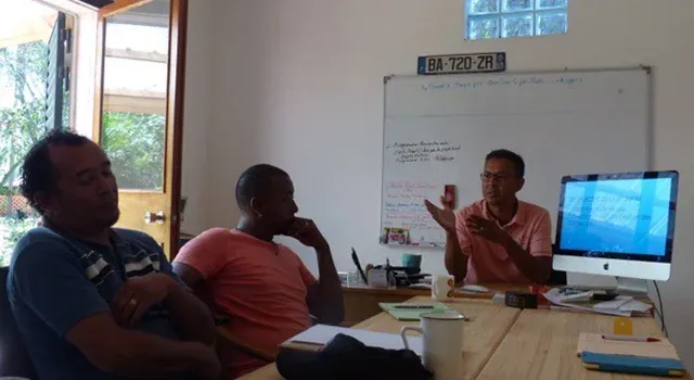 Réunion en salle avec trois participants discutant autour d’une table et d’un tableau blanc avec notes