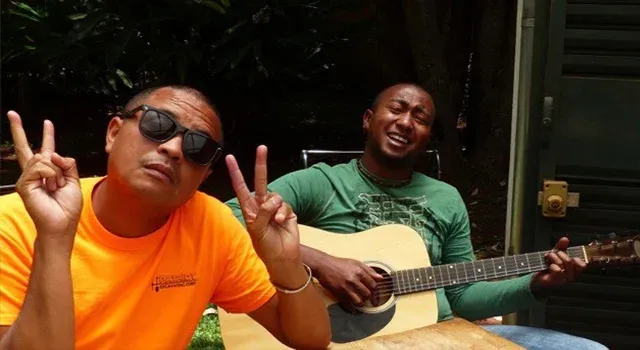 Deux hommes souriants en extérieur l’un faisant des signes de paix l’autre jouant de la guitare acoustique
