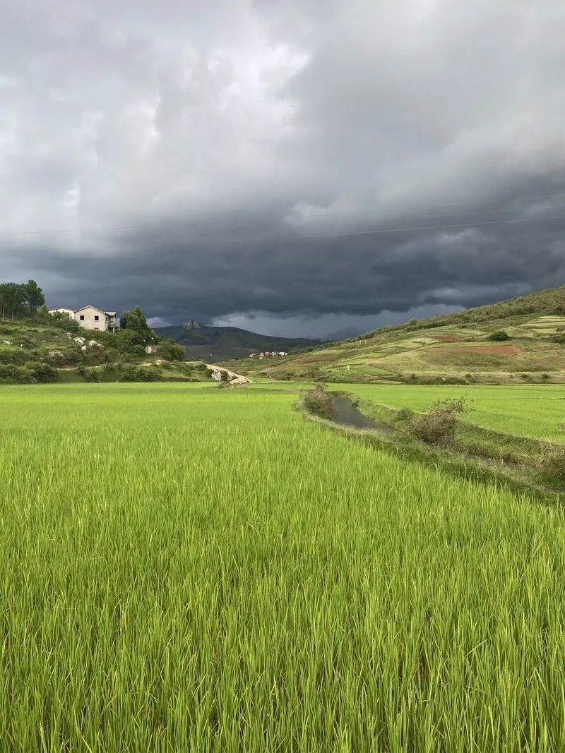 Champ verdoyant sous orage menaçant avec maisons en hauteur et montagnes lointaines